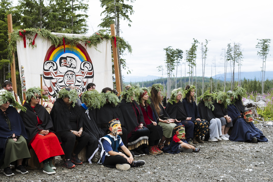 Mourning ceremony in clearcut, a row of women mourners wearing hemlock boughs, children wearing carved mourning masks sitting in front