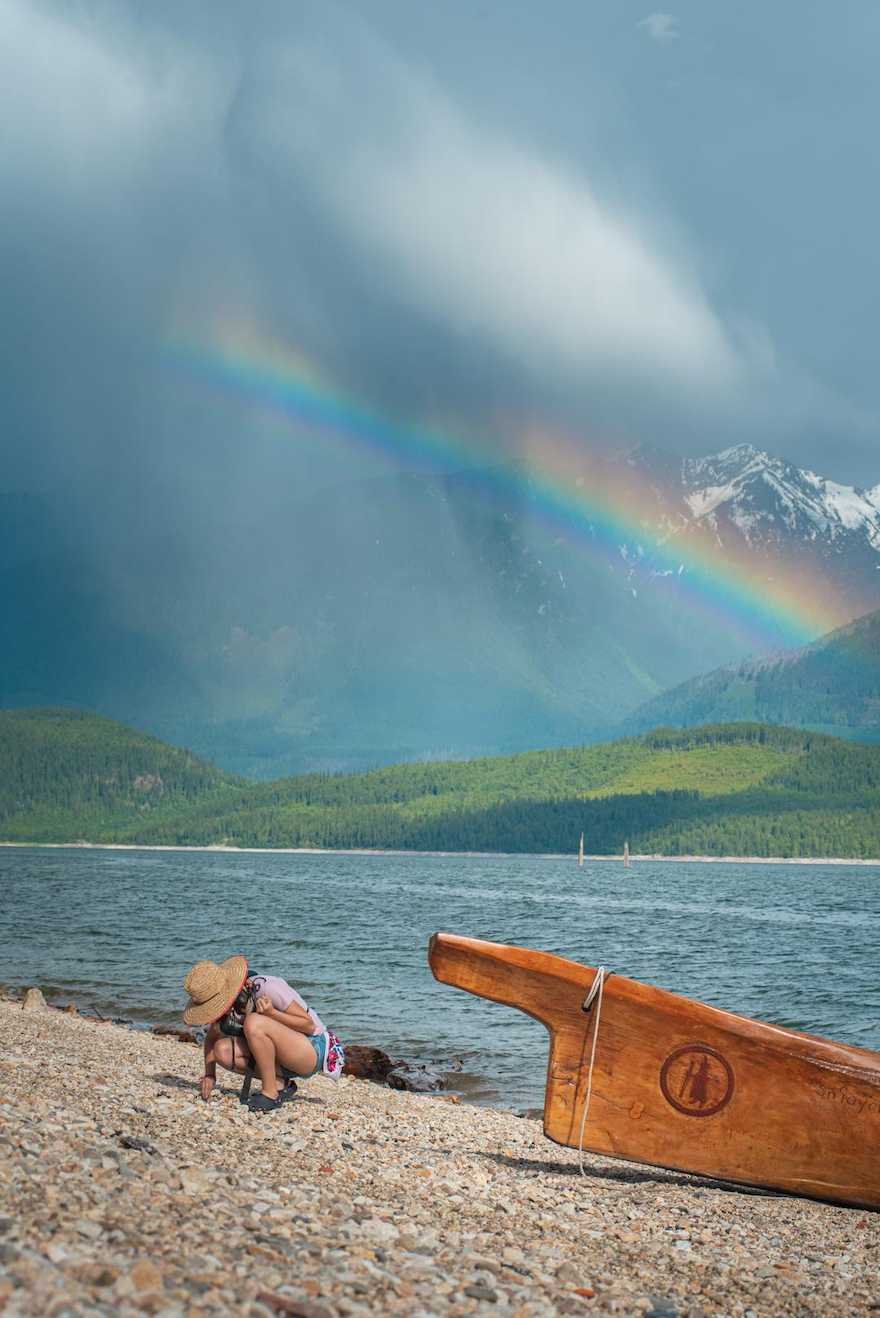 Sinixt woman on a pebble beach with a canoe, mountains and a rainbow behind