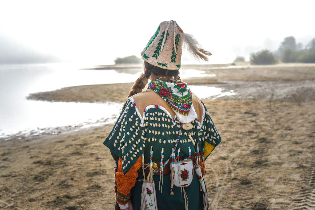 Sinixt Elder and cultural leader Shelly Boyd in regalia, standing on a beach facing the water