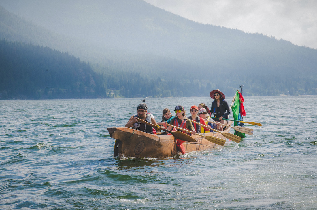 Sinixt people paddling a canoe together on a lake with mountains behind
