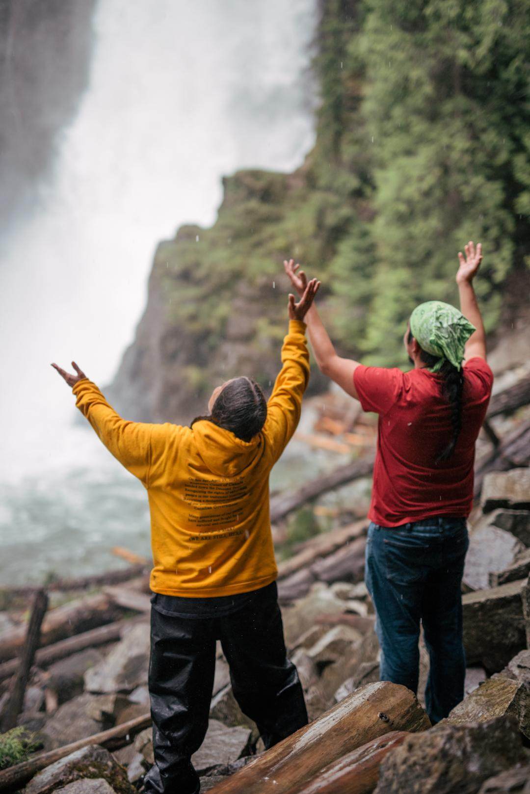 Two Sinixt people standing in front of waterfall with raised arms