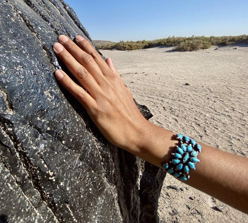 Hand with turquoise bracelet resting on obsidian rock outcropping, sand and sky in background